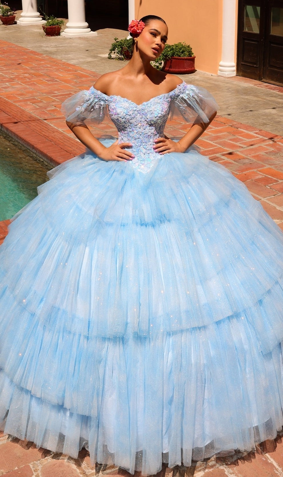 A woman stands by a brick walkway and pool, wearing Quinceanera Dress 54312 By Amarra?a light blue, off-the-shoulder ball gown?with a red flower in her hair.